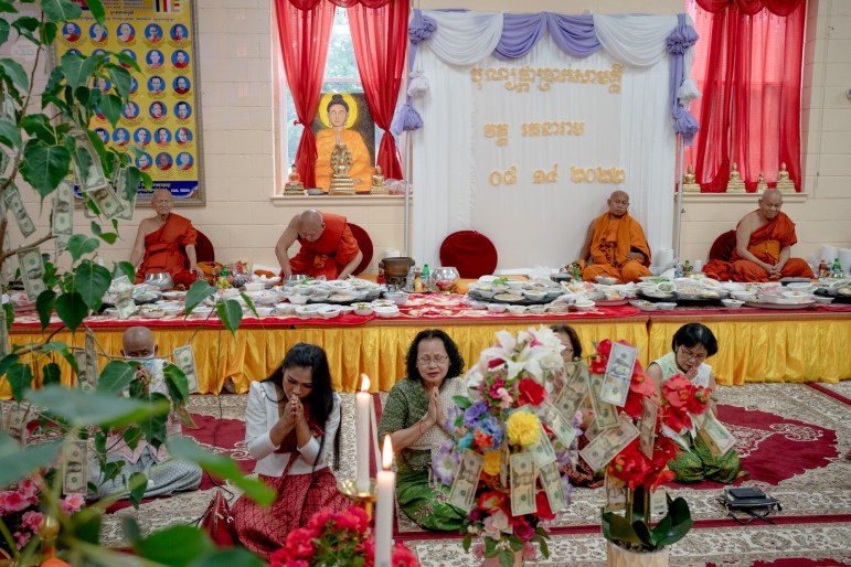 A row of five people are seated on a carpet with hands in a prayer position. Behind them, a row of four monks sit at a table.