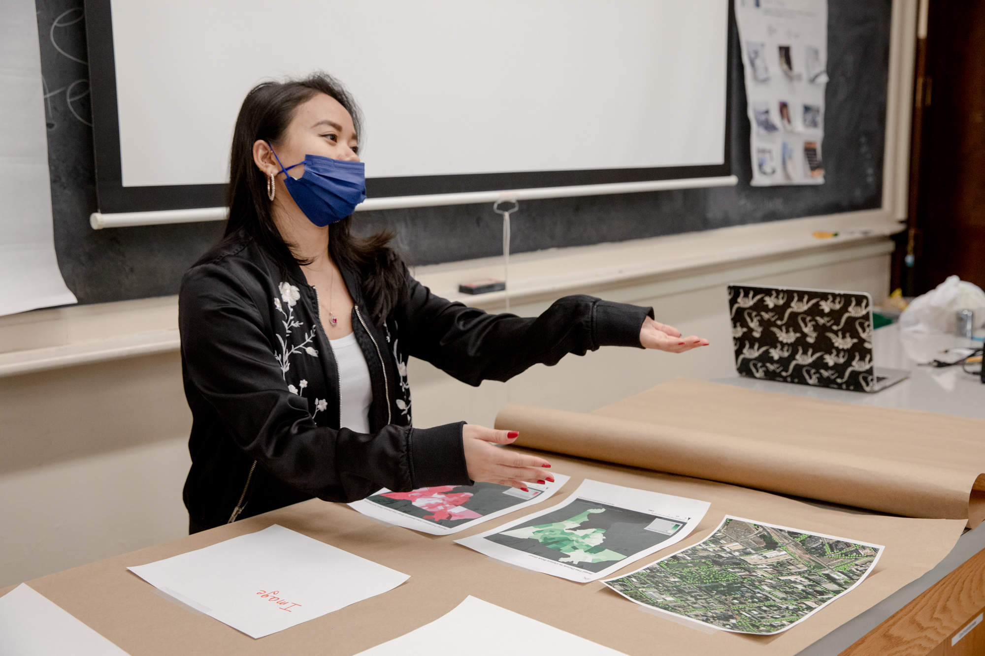 Natasha Ghazali sits at a table with her hands outstretched. She is wearing a mask and a jacket.