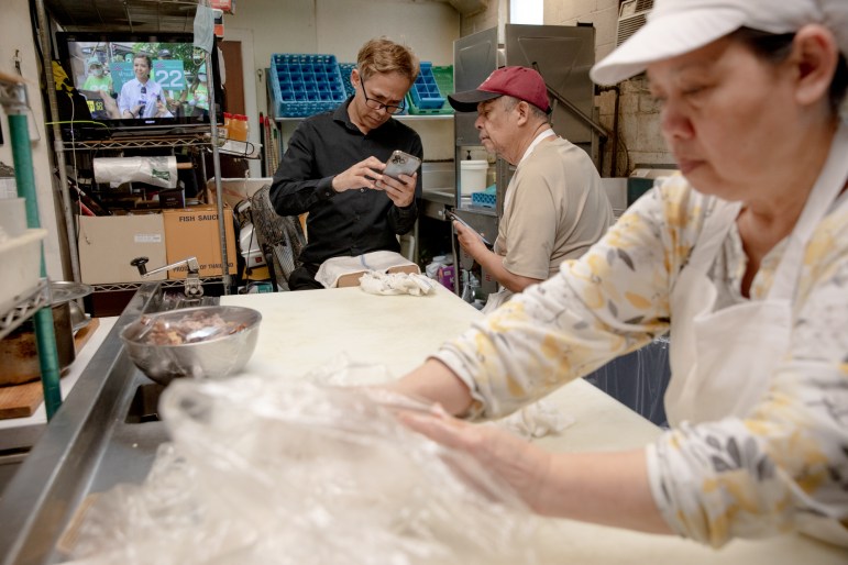 Theeradej Juntorn looks at his phone in the kitchen of a restaurant. Another man is looking at the phone as well. A woman is preparing food at a counter.