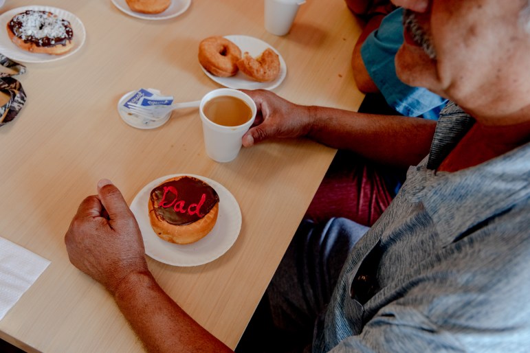 A man sits at a table with a cup of coffee and several plates with doughnuts. One of the doughnuts has the word "Dad" written on it.