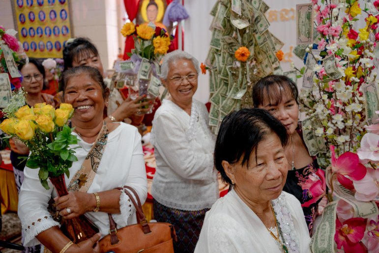A group of women in procession smile while holding flowers and towers of dollar bills.