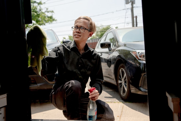 Theeradej Juntorn is knelt down, wiping glass with a rag in one hand and holding a bottle of Windex in the other.