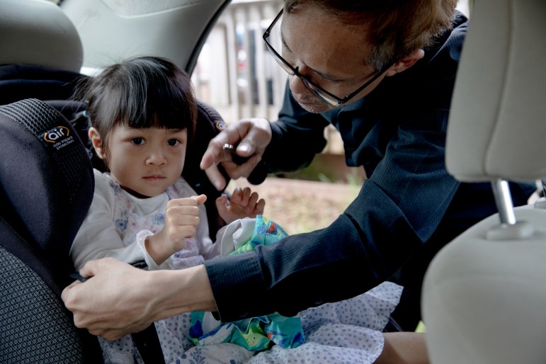 Theeradej Juntorn loads his daughter Kyly into a carseat. Kyly is wearing a dress with dots on it.