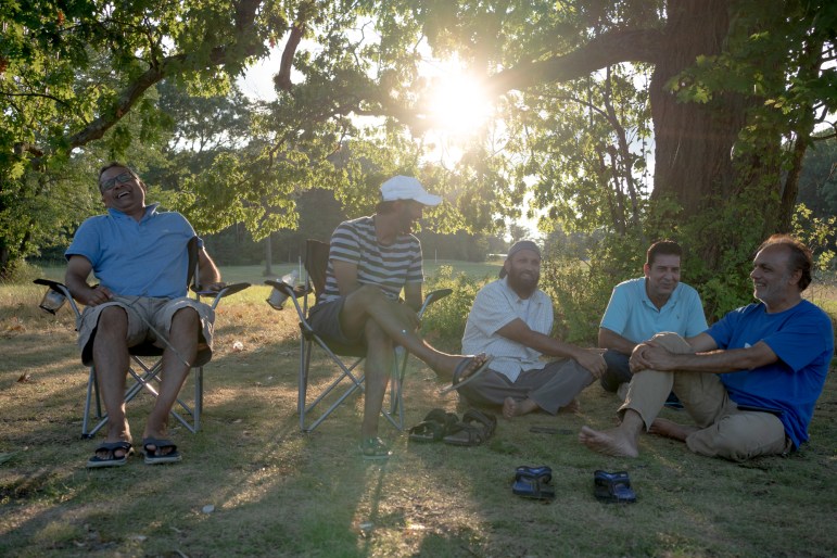 Five men sit laughing near a large tree. The sun shines through the leaves.