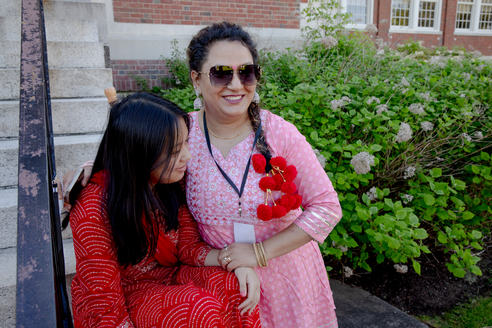 Chandani Khatri Chhetri stands and hugs Namrata Malla, who is seated. They are outdoors standing next to a building.
