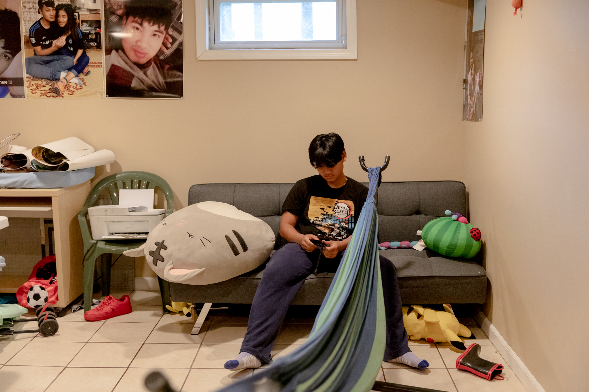 Jet Smee sits on a couch looking at his cell phone. He is wearing a t-shirt, sweatpants, and socks. There are a few stuffed animals on and underneath the couch.