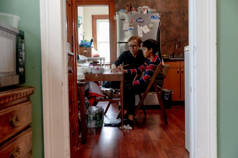 Theeradej Juntorn and his son Caney, sit at a table in the kitchen. Theeradej is pointing at the homework in front of them. Caney is holding a pencil in his right hand.