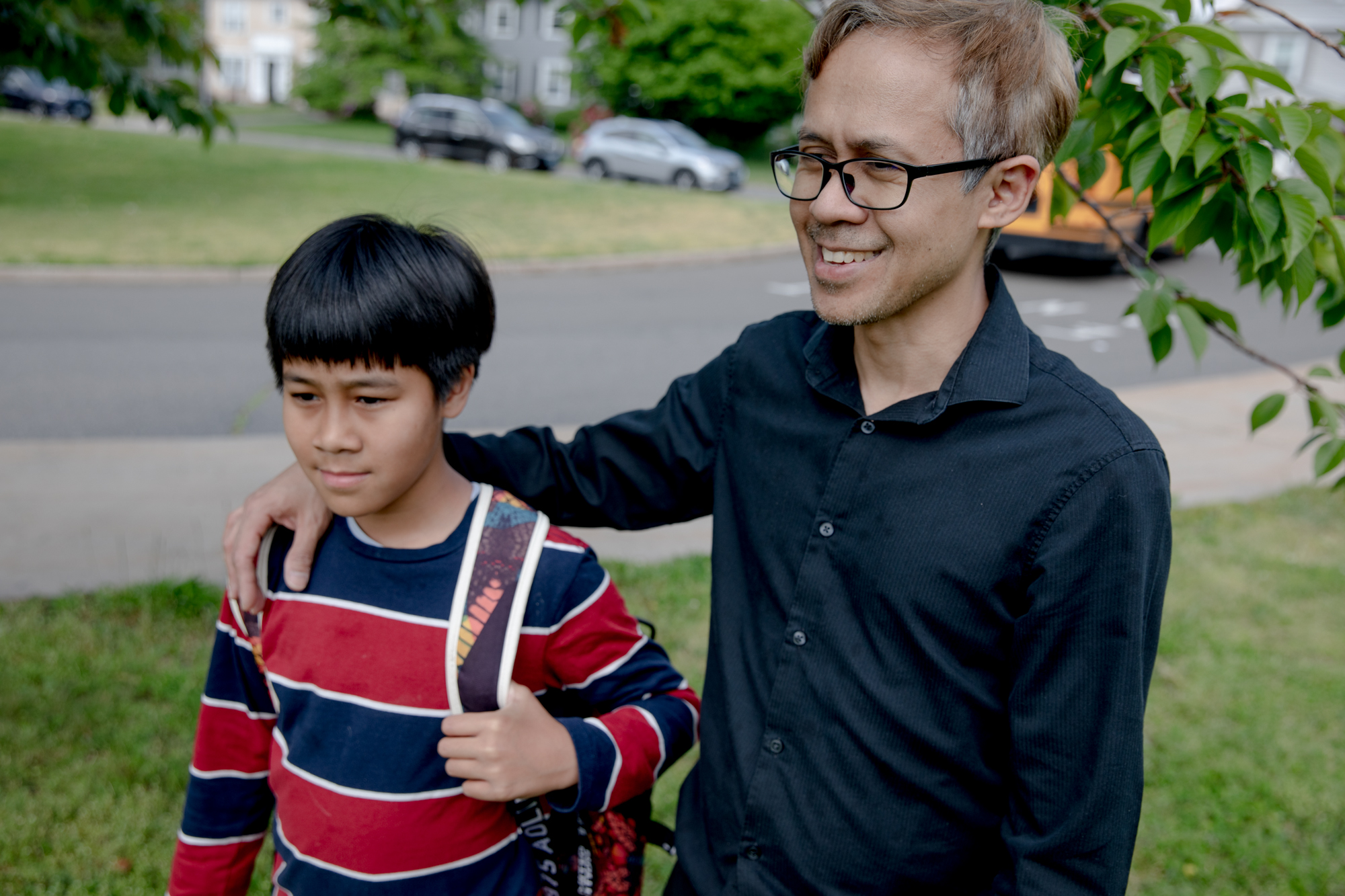 Theeradej Juntorn stands with one arm around his son, Caney. Theeradej is wearing a collared shirt and glasses. Caney is wearing a long sleeved t-shirt and has a backpack slung over one shoulder.