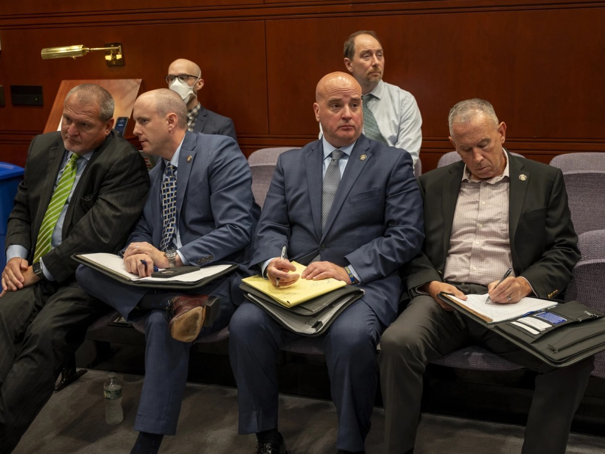 Connecticut State Police Union representative Andrew Matthews sits in a room in Hartford's Legislative Office Building. He's hold a note pad and pen. He's sitting next to other union representatives.