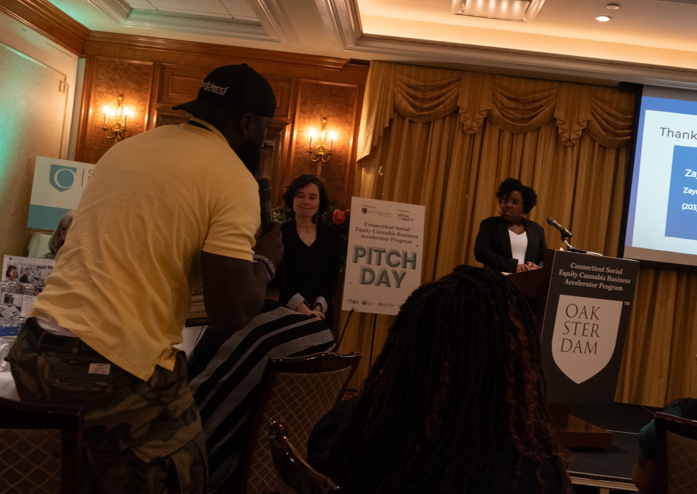 A woman at a podium answers a question from a man holding a mic in the audience.