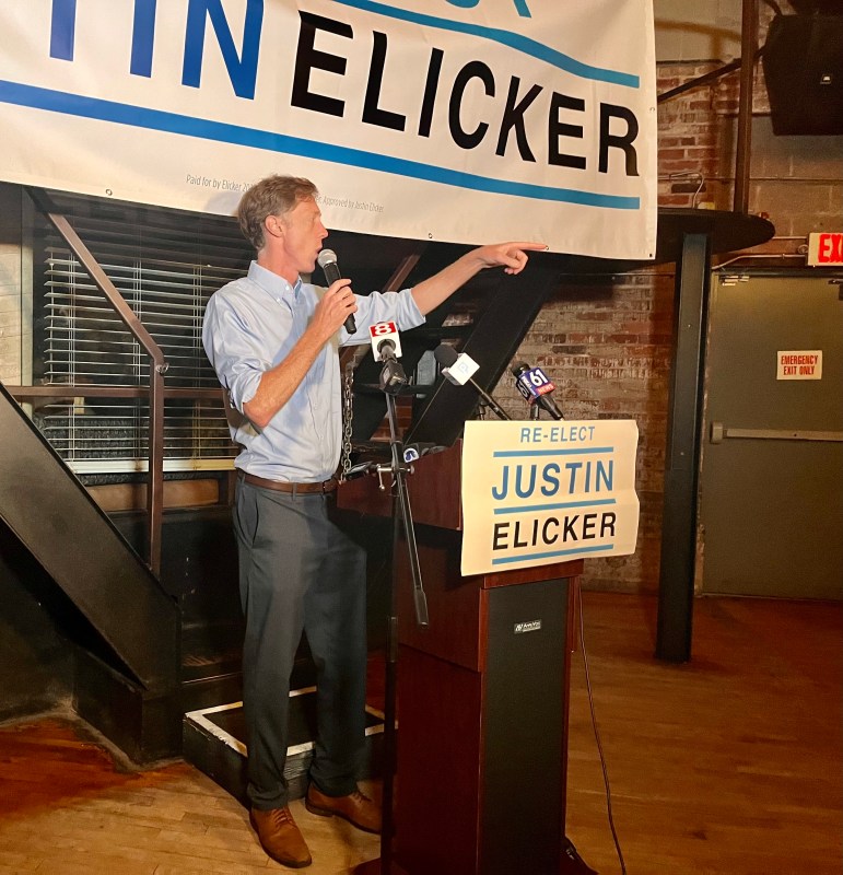 New Haven Mayor Justin Elicker speaks at a lectern with a Re-Elect Justin Elicker sign in front of him.