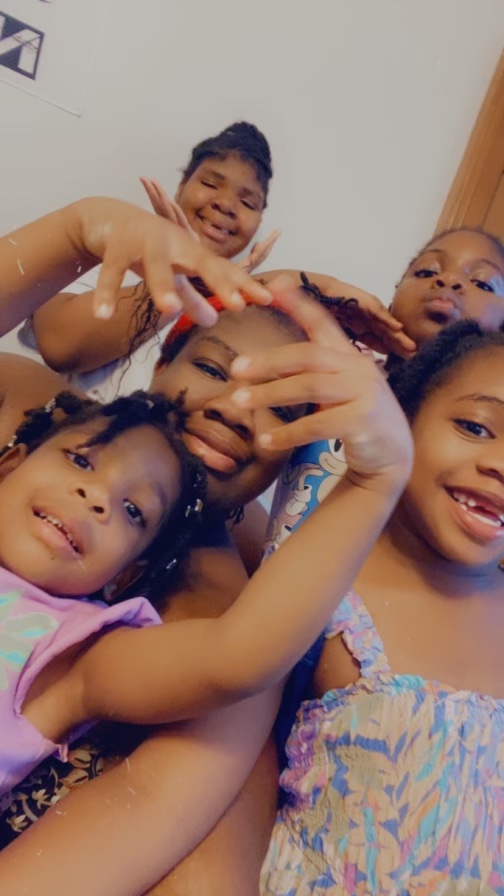 A woman and her four young daughters pose for a selfie. One of the girls in the front has her hands above her head.