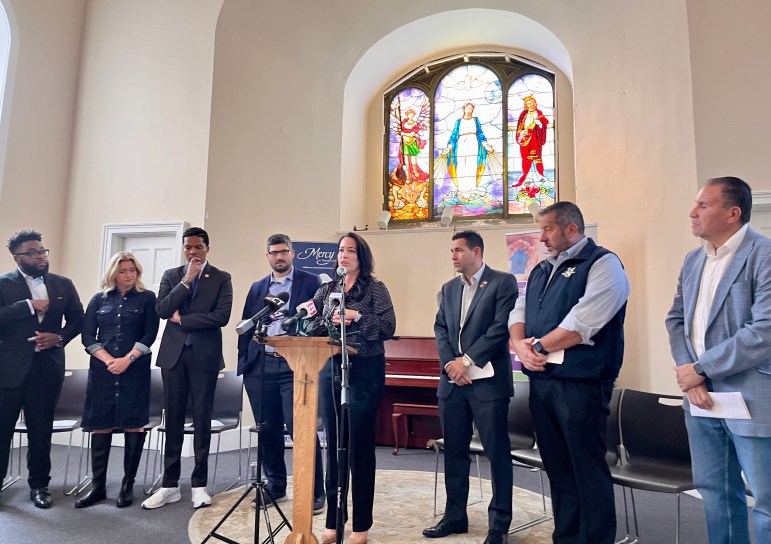 A woman at a lecturn speaks into microphones, with state lawmakers on both sides of her.