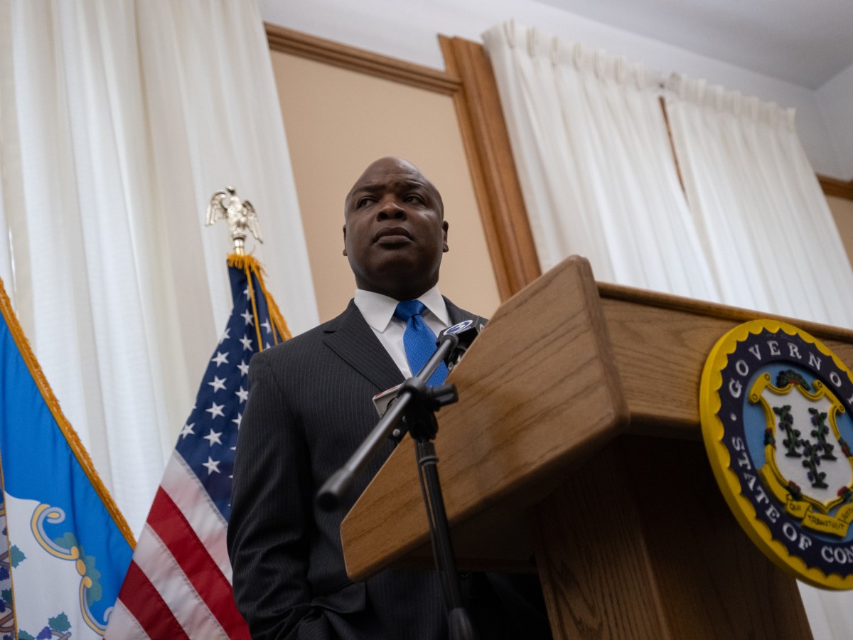 Ronnell Higgins stands behind a podium in a conference room at the State Capitol. He is listening to questions from members of the press.