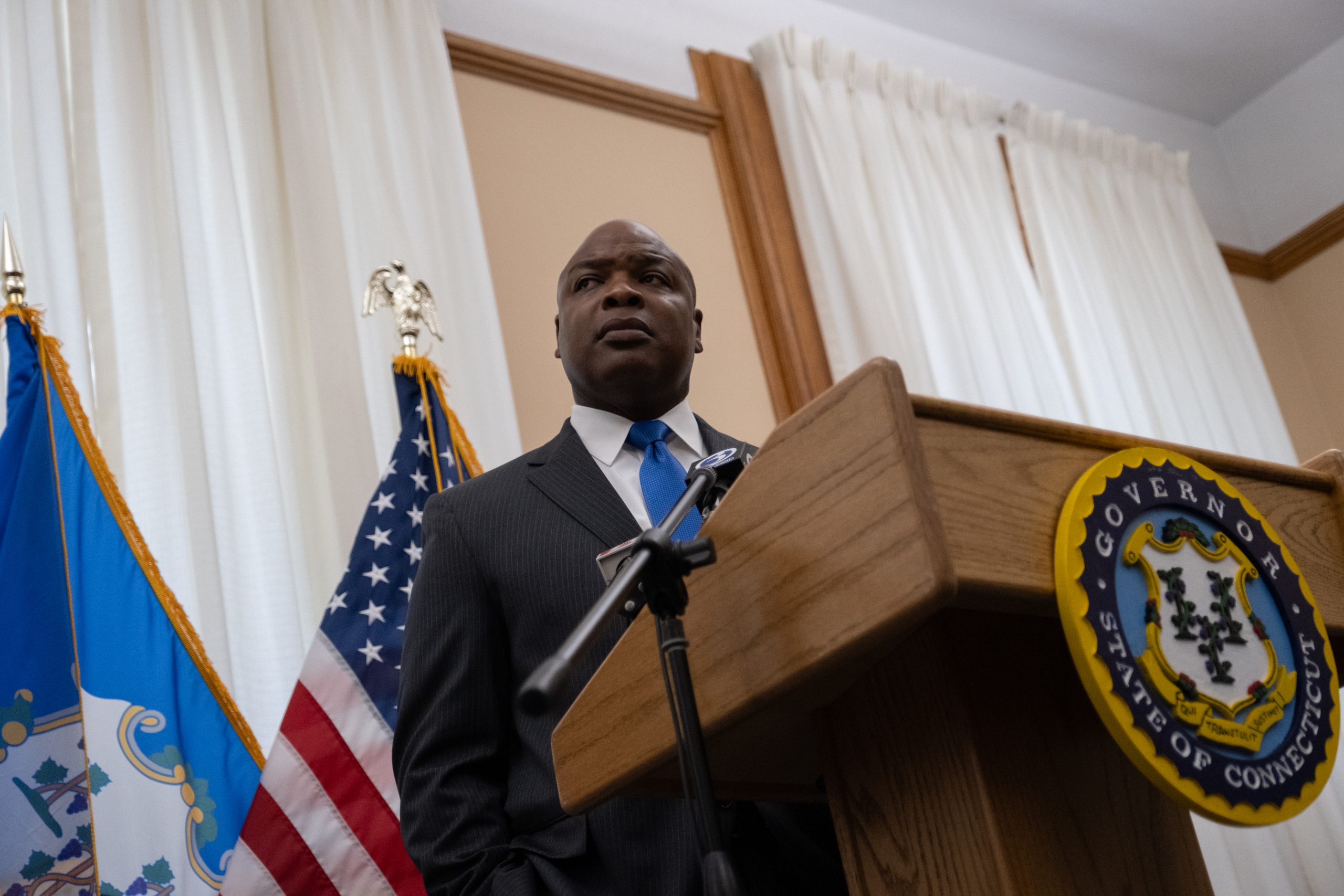 Ronnell Higgins stands behind a podium in a conference room at the State Capitol. He is listening to questions from members of the press.