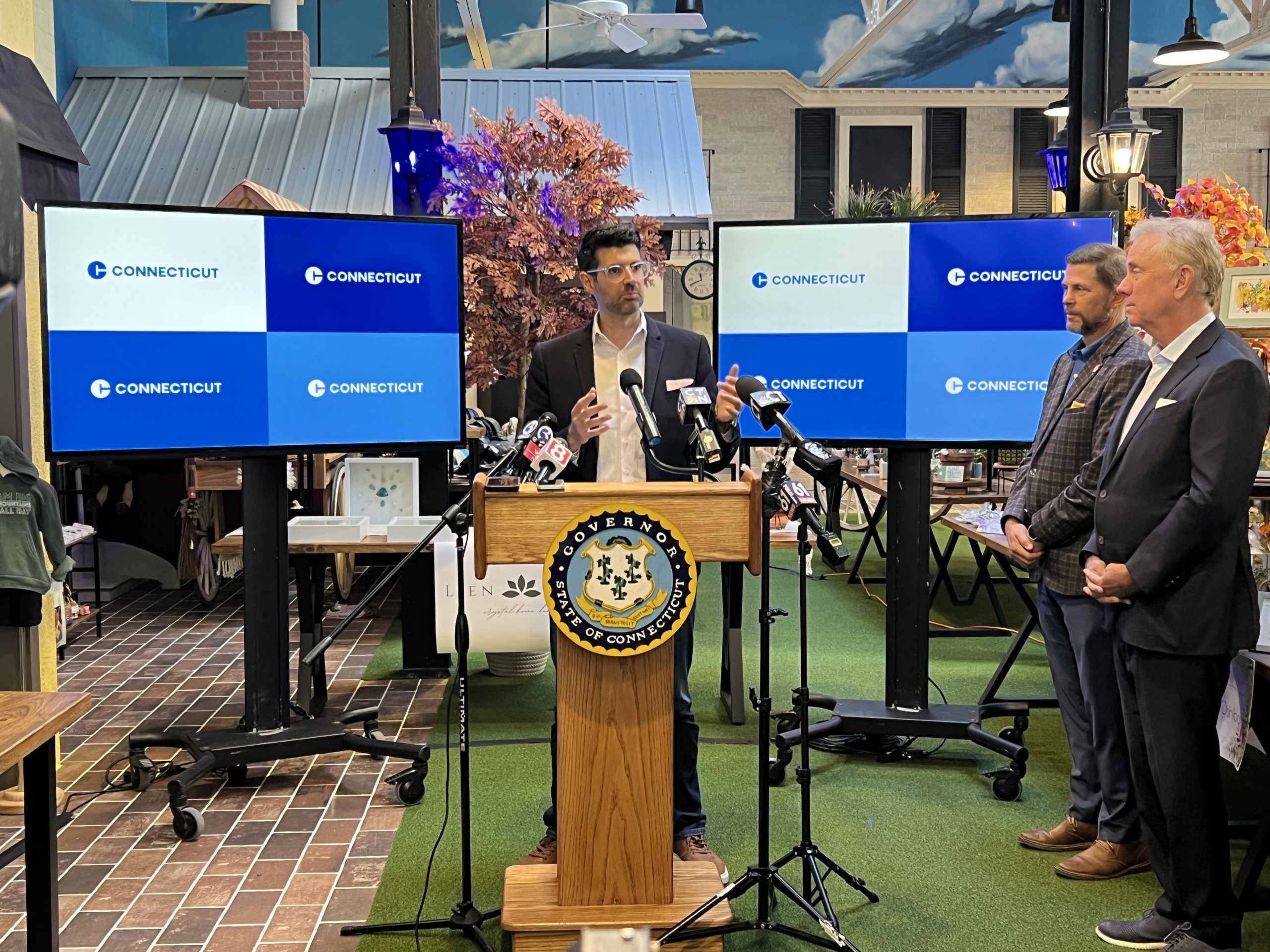 A man stands at a podium with TV screens behind him, showing Connecticut's new marketing logo.