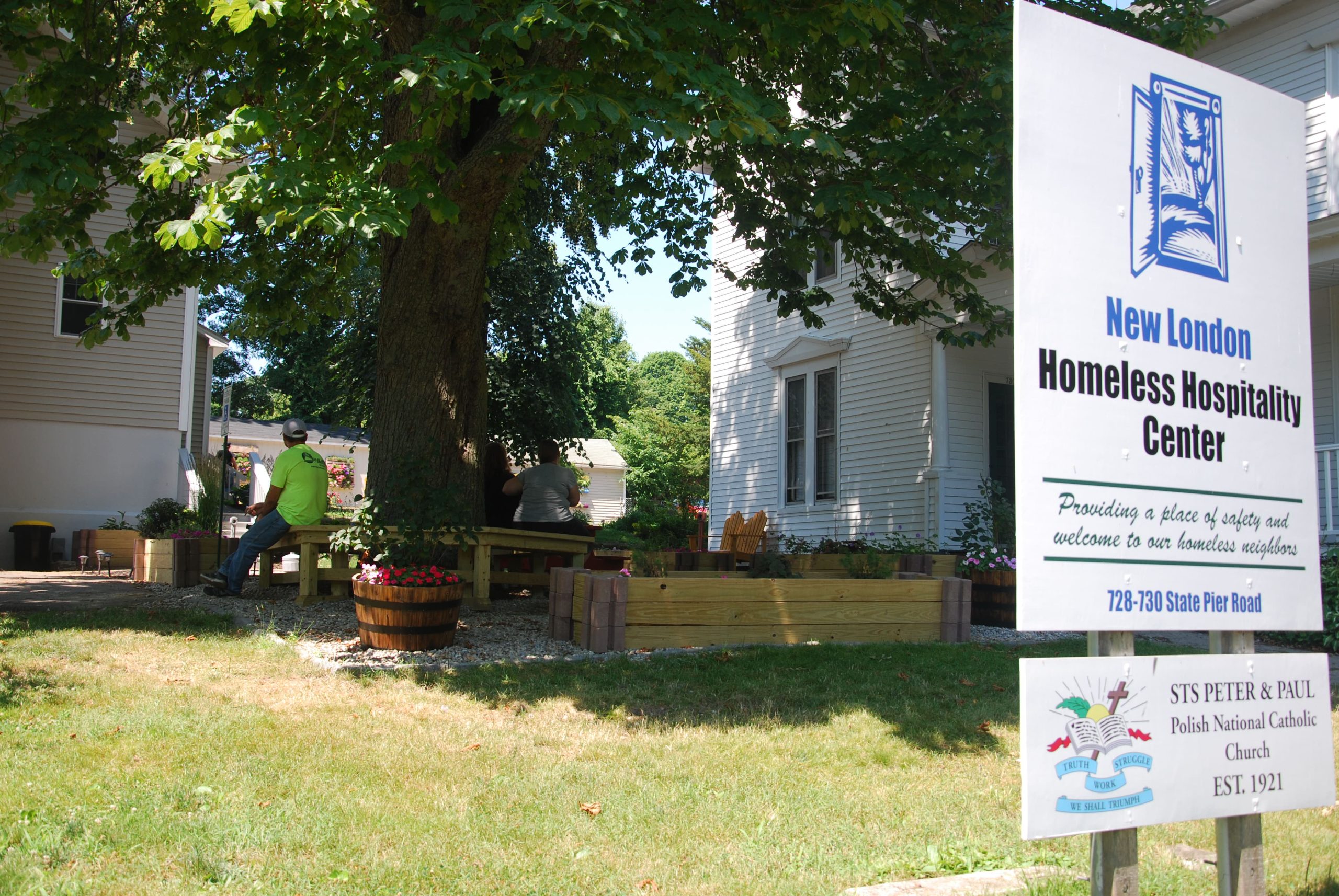 A sign for the New London Homeless Hospitality Center is in the foreground, with a tree and two buildings in the background.