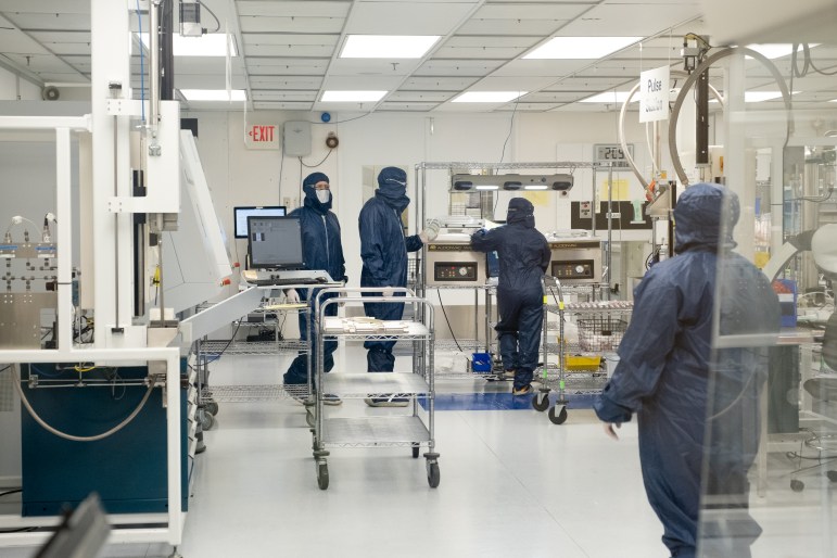 Engineers in coveralls and hoods operate machines in a brightly-lit room.