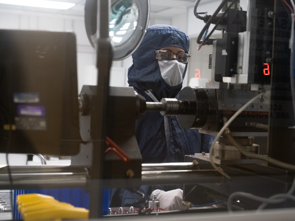 A worker in hooded, full-body coveralls and goggles works at a welding machine behind double-paned glass.