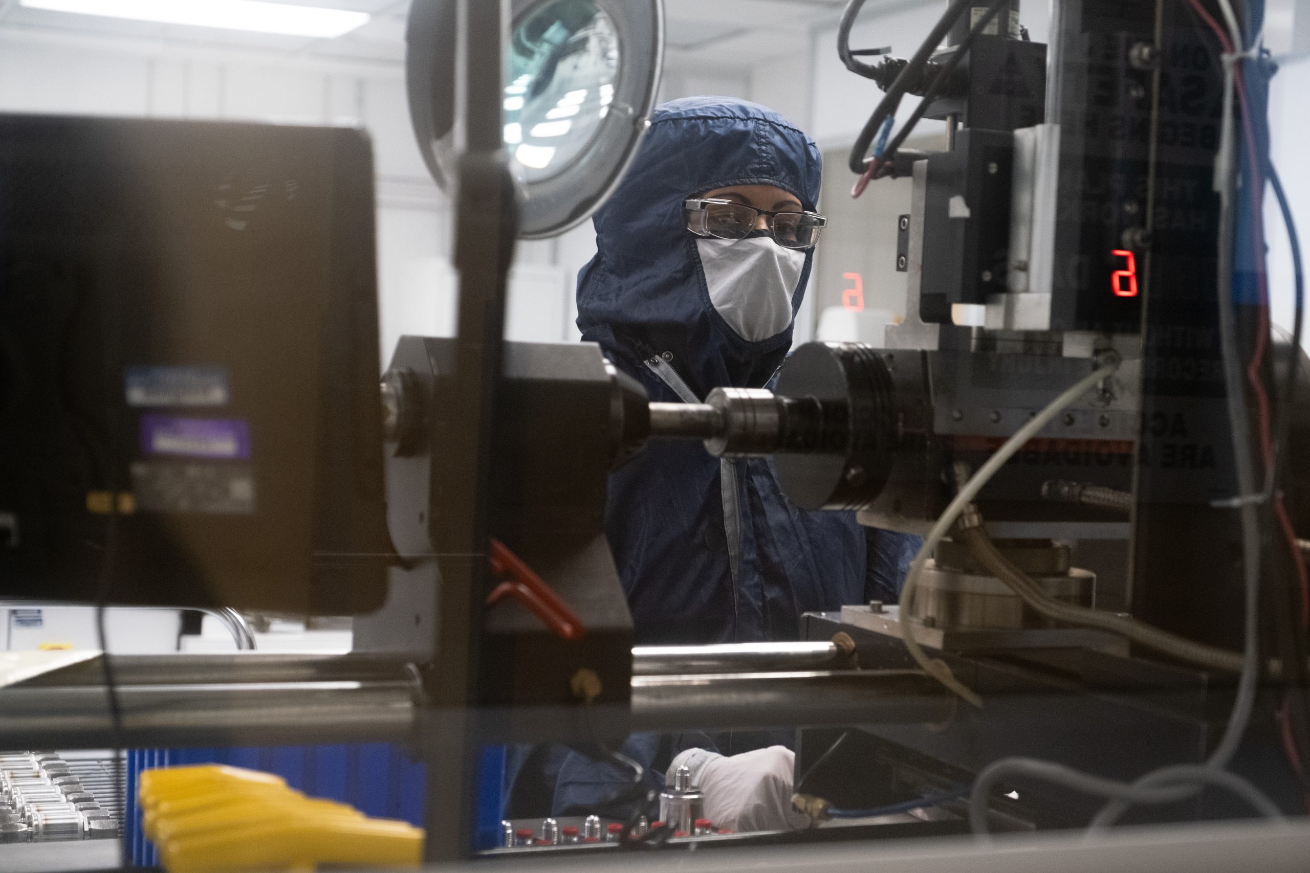 A worker in hooded, full-body coveralls and goggles works at a welding machine behind double-paned glass.