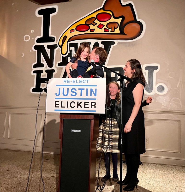 New Haven Mayor Justin Elicker stands behind a lecturn as he claims victory in the 2023 General Election. He's holding his daughter -- a young girl while his daughter and wife stand beside him.