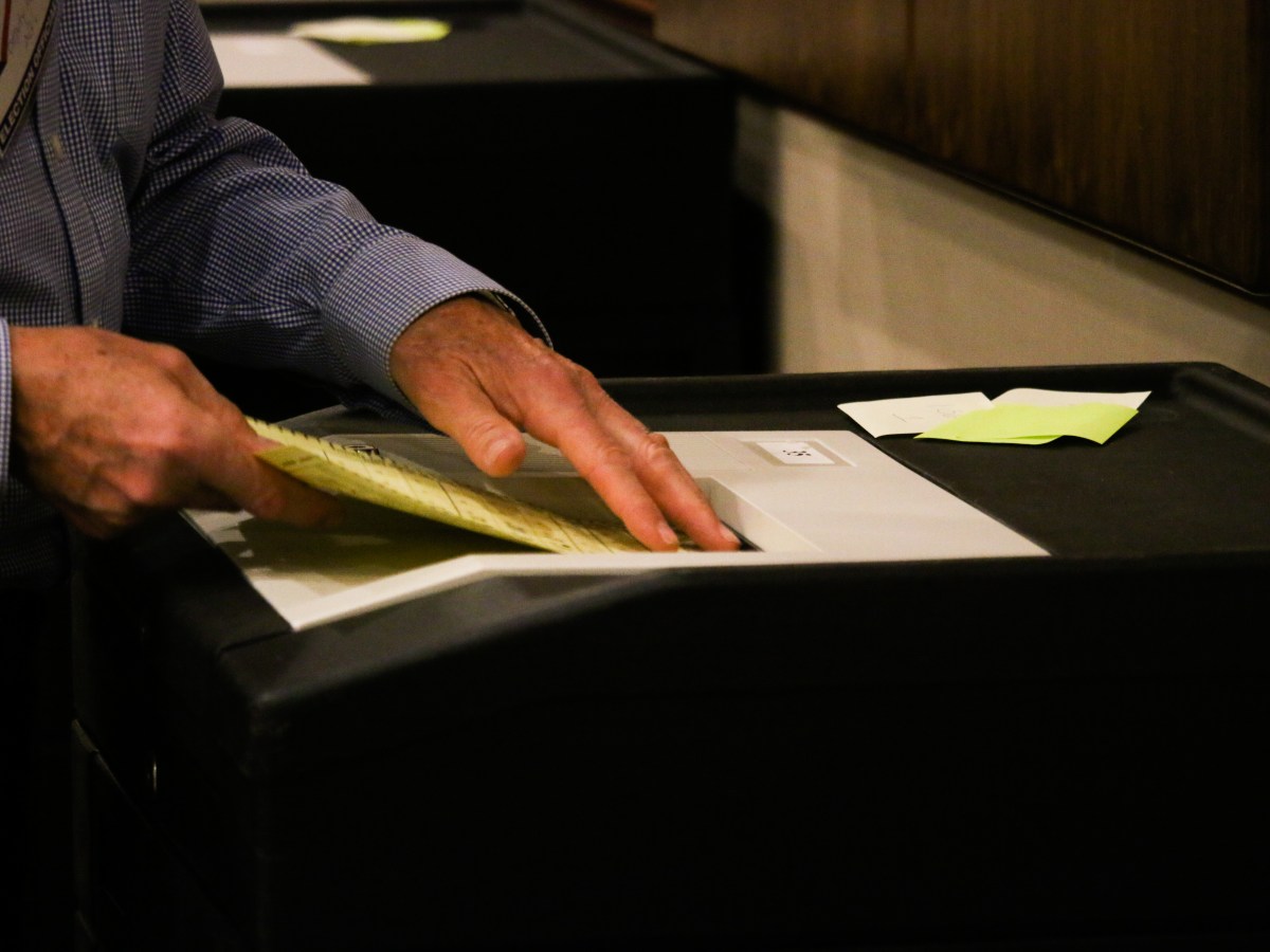 Hands are seen inserting an election ballot — a piece of yellow paper — into a machine that calculates vote totals.