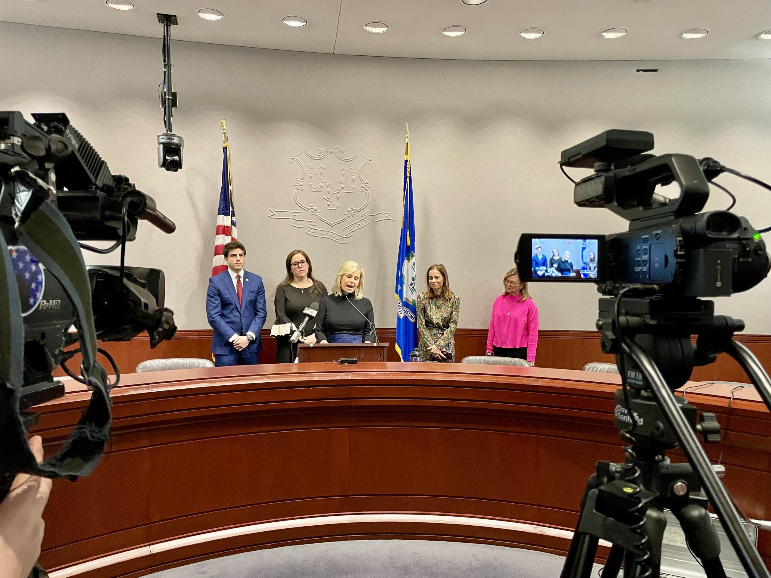 A woman speaks at a podium with TV cameras in the foreground.