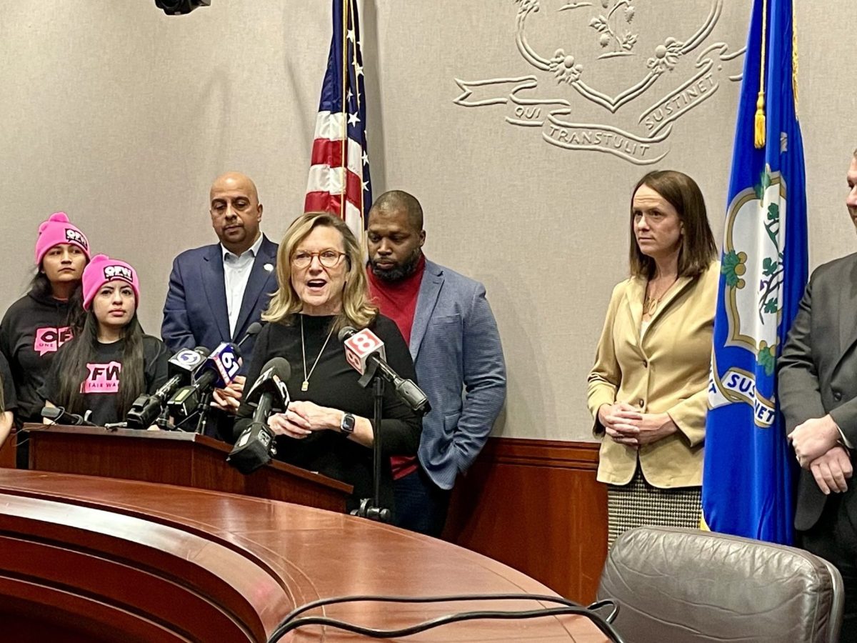 A woman at a podium, flanked by supporters wearing 'One Fair Wage' hats.