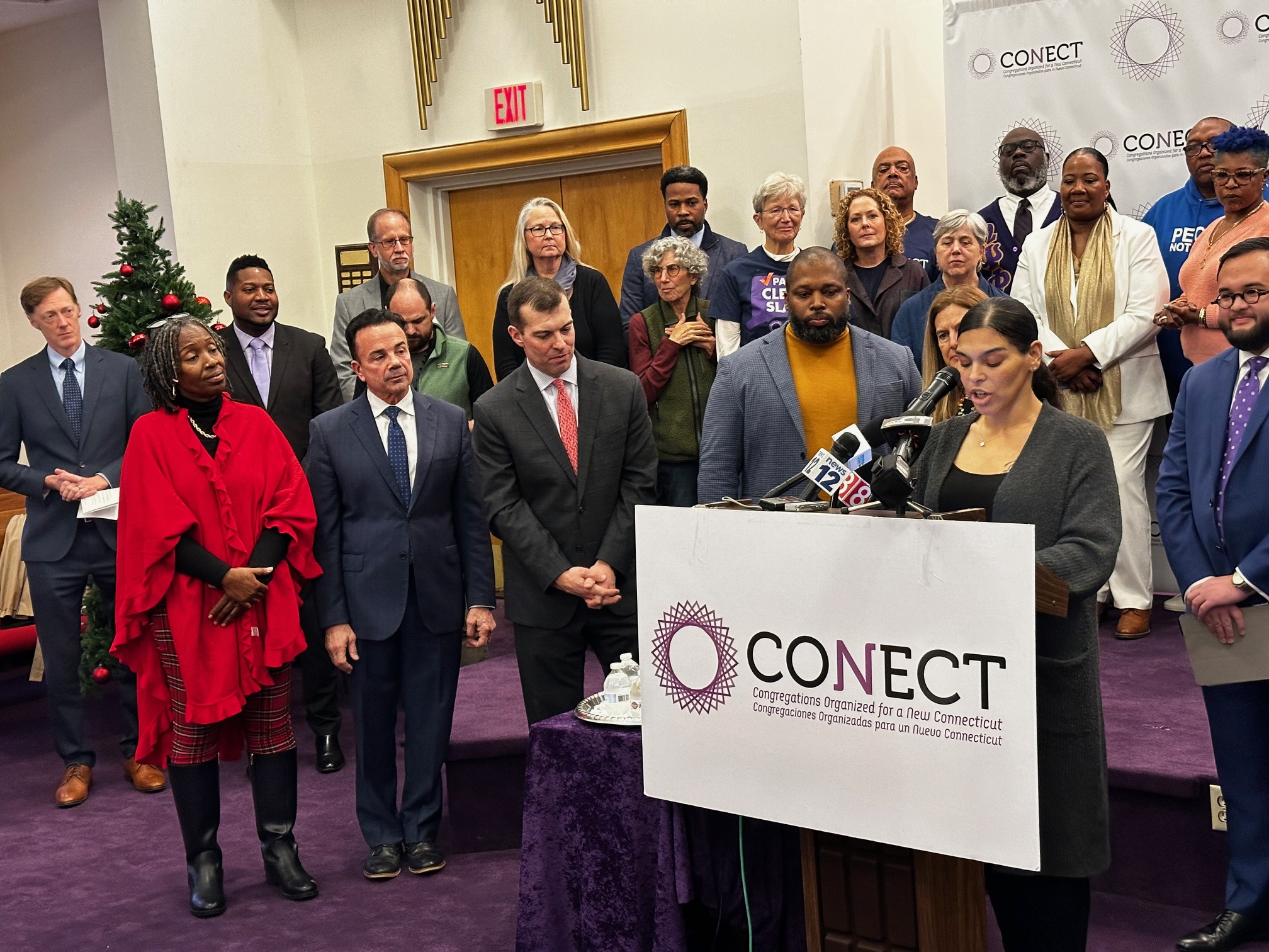 A group of Connecticut residents, lawmakers and advocates stand behind and on the side a person speaking at podium inside of a New Haven church. All of the people are at a gathering to announce the full rollout of Connecticut's "clean slate" law, which automatically erases misdemeanors and low-level felonies from the criminal records of some people.