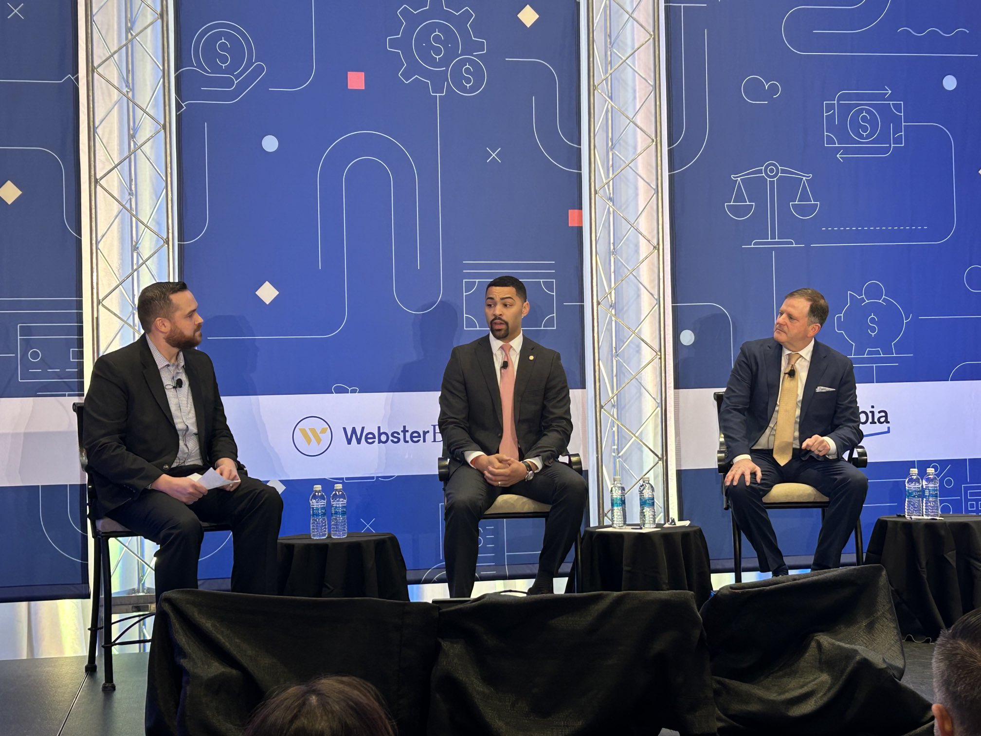 Three men speak on a panel on a stage with a blue background.