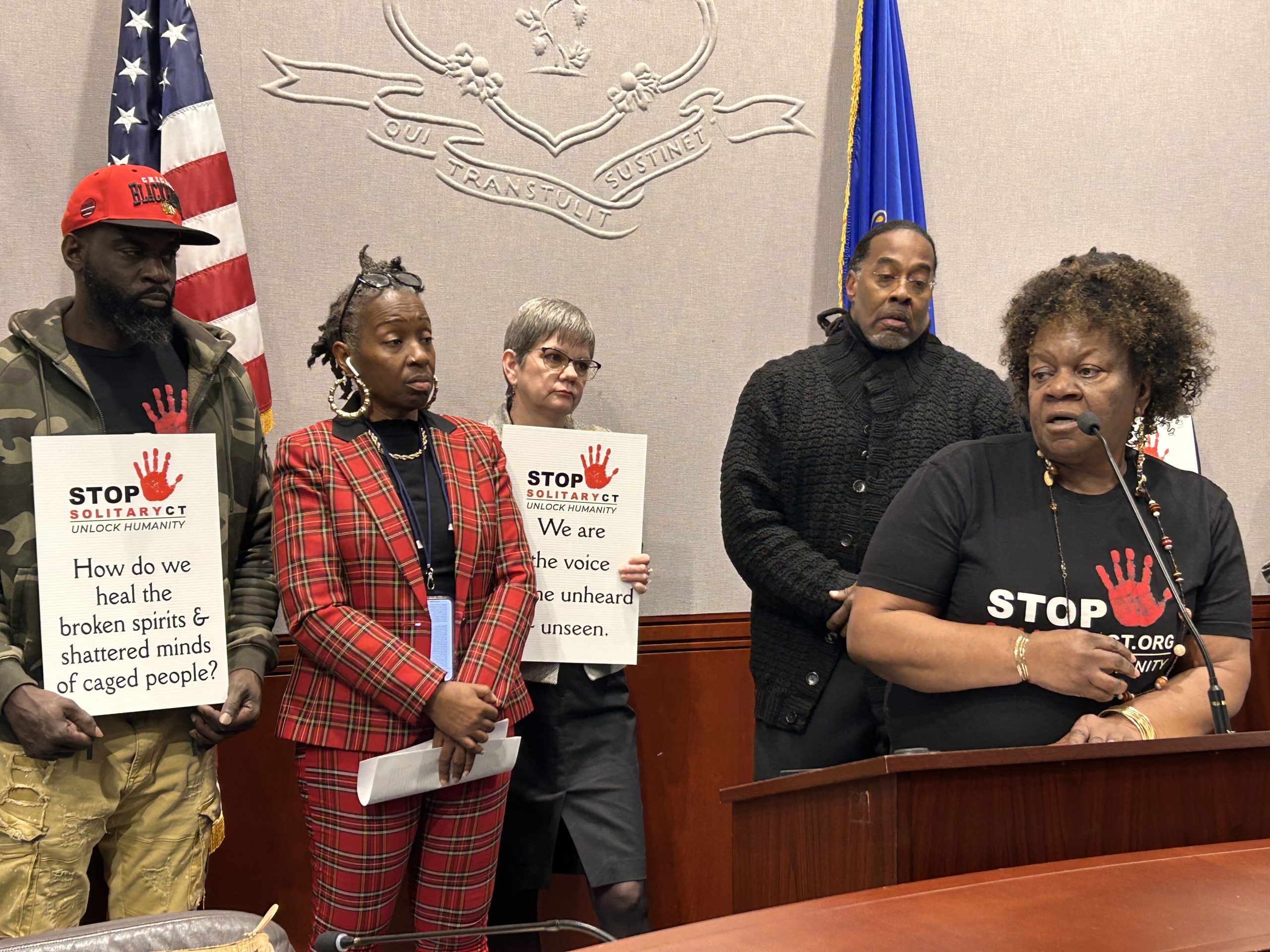 Barbara Fair, the leading organizer for Stop Solitary CT, is standing at a podium and speaking into a microphone at the Legislative Office Building in Hartford. Lawmakers and advocates are standing behind her holding signs with the organization's logo and statements in support of incarcerated people.