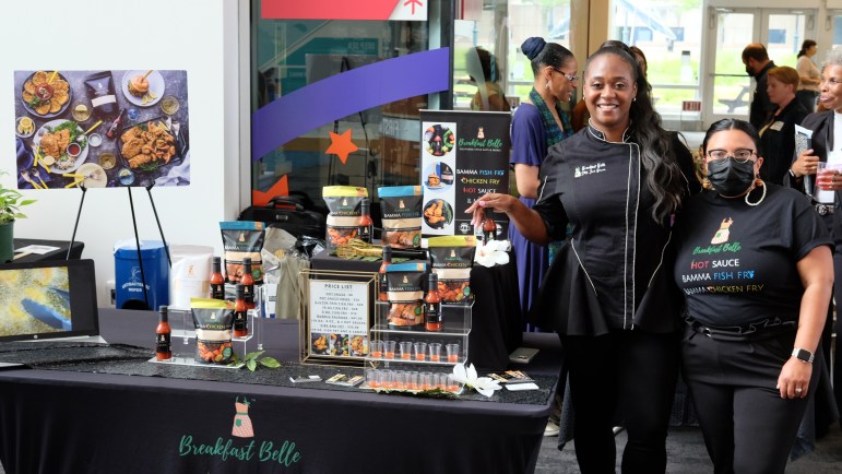 Two women stand beside a table displaying hot sauces, packs of breading mix and photos of food.