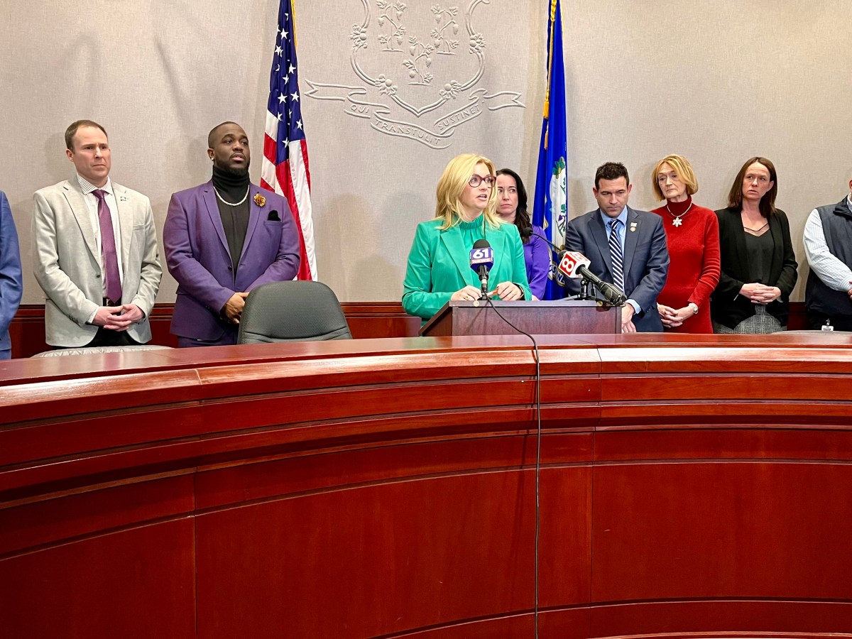 A state representative speaks at a lecturn in front of the American and Connecticut flags. She is surrounded by other lawmakers.