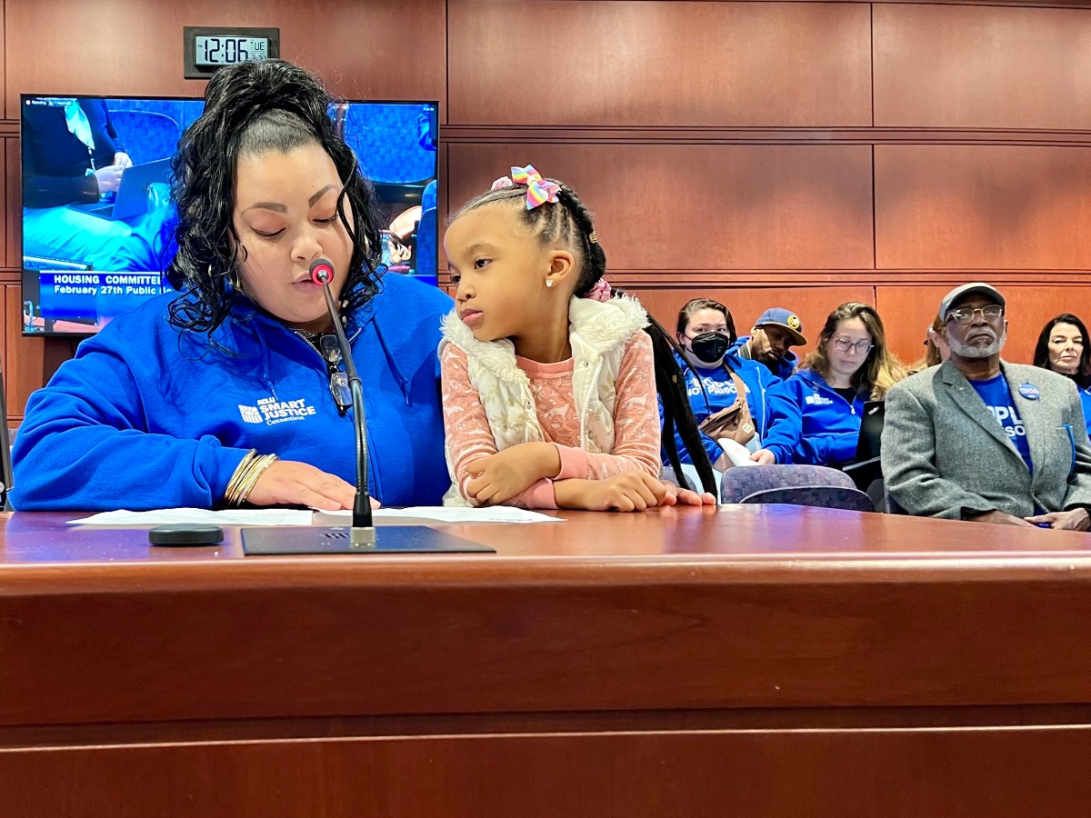 A woman sits in front of a microphone. People in ACLU shirts are behind her and a 4-year-old girl sits in her lap.