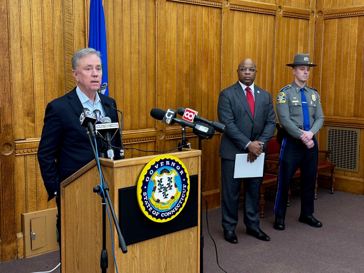 Gov. Ned Lamont is standing behind a podium and speaking to members of the press. Two State Police officials are standing to his right, one of whom is Ronnell Higgins, the commissioner of the Department of Emergency Services and Public Protection.