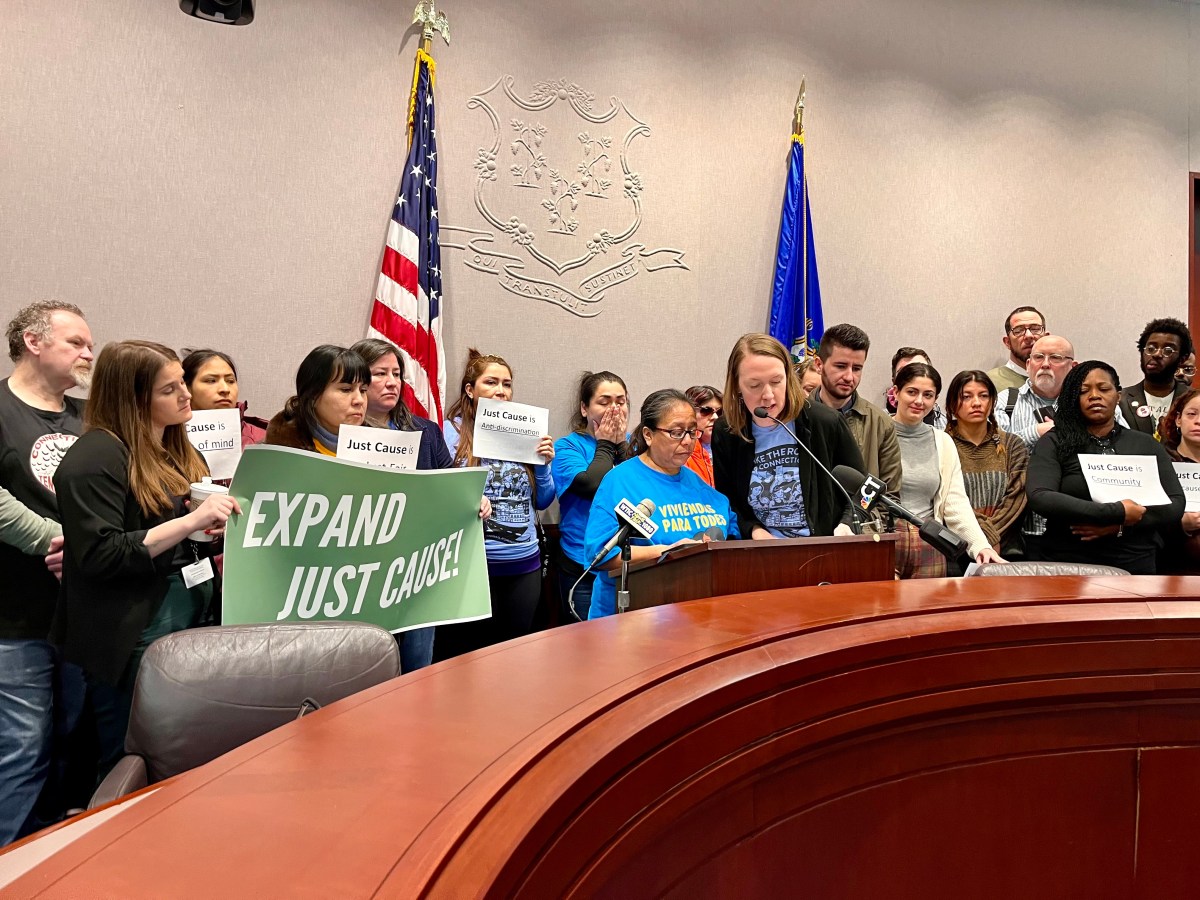 Two women are gathered at a lecturn. They are wearing blue shirts, and people gathered around them are holding signs calling for an expansion of Just Cause eviction protections