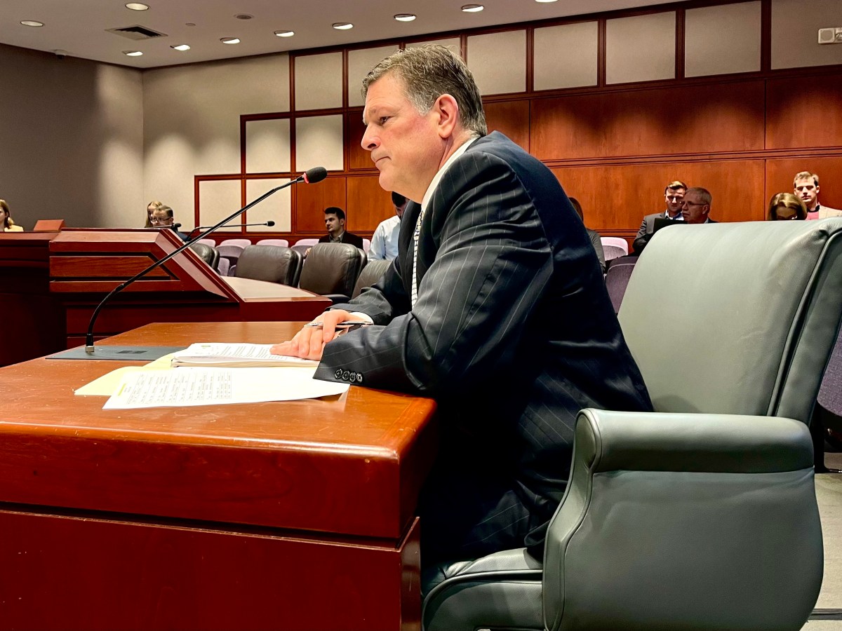 A man in a suit sits at a desk with a microphone and papers in front of him.