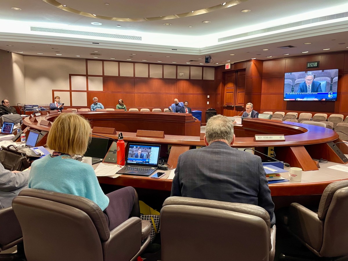 Lawmakers seated around a table in a hearing room, listening to a male speaker.