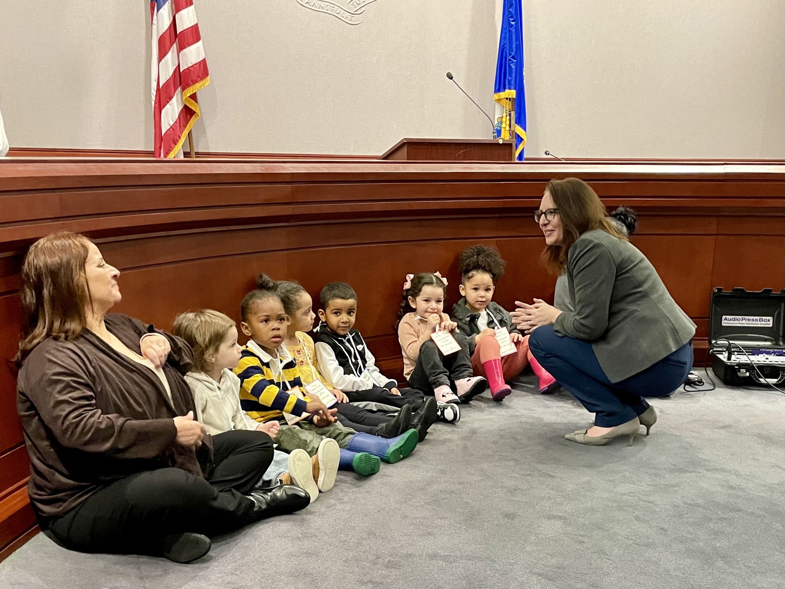 A woman kneels down to speak with toddlers, who are seated in front of a dais in the Legislative Office Building.