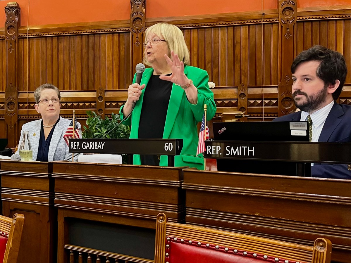A woman in a green blazer stands on the House floor holding a microphone. Another lawmaker looks up at her while she describes the bill.