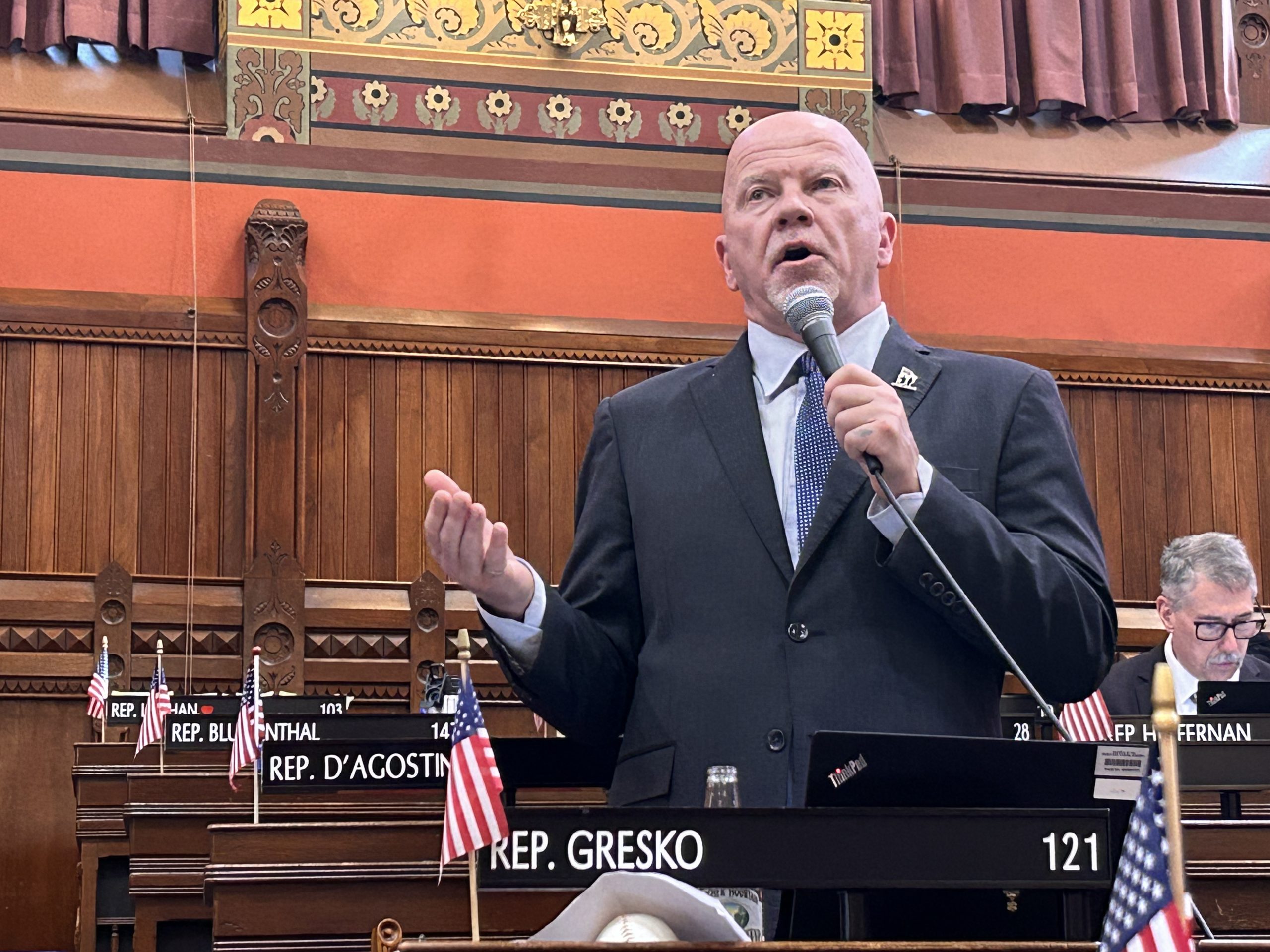 A man in a suit stands with a microphone on the House floor.