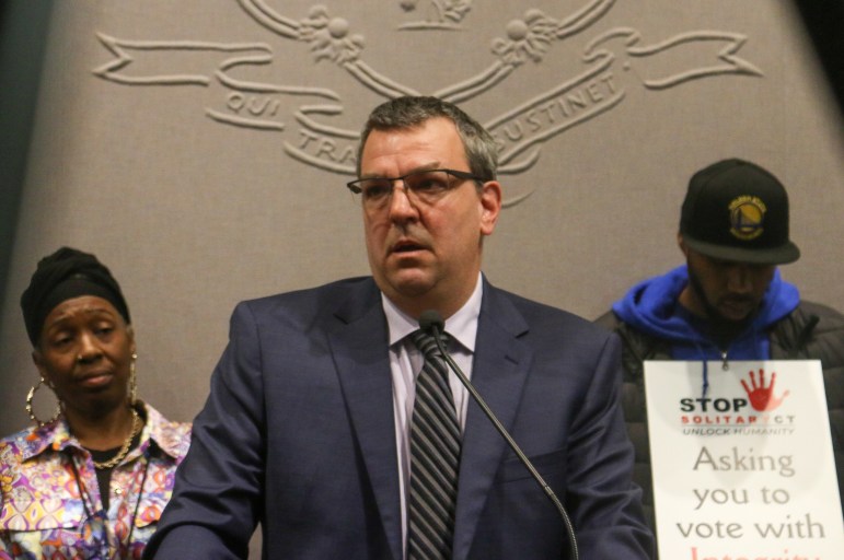 Ken Krayeske is speaking into a microphone while two people are standing behind him.