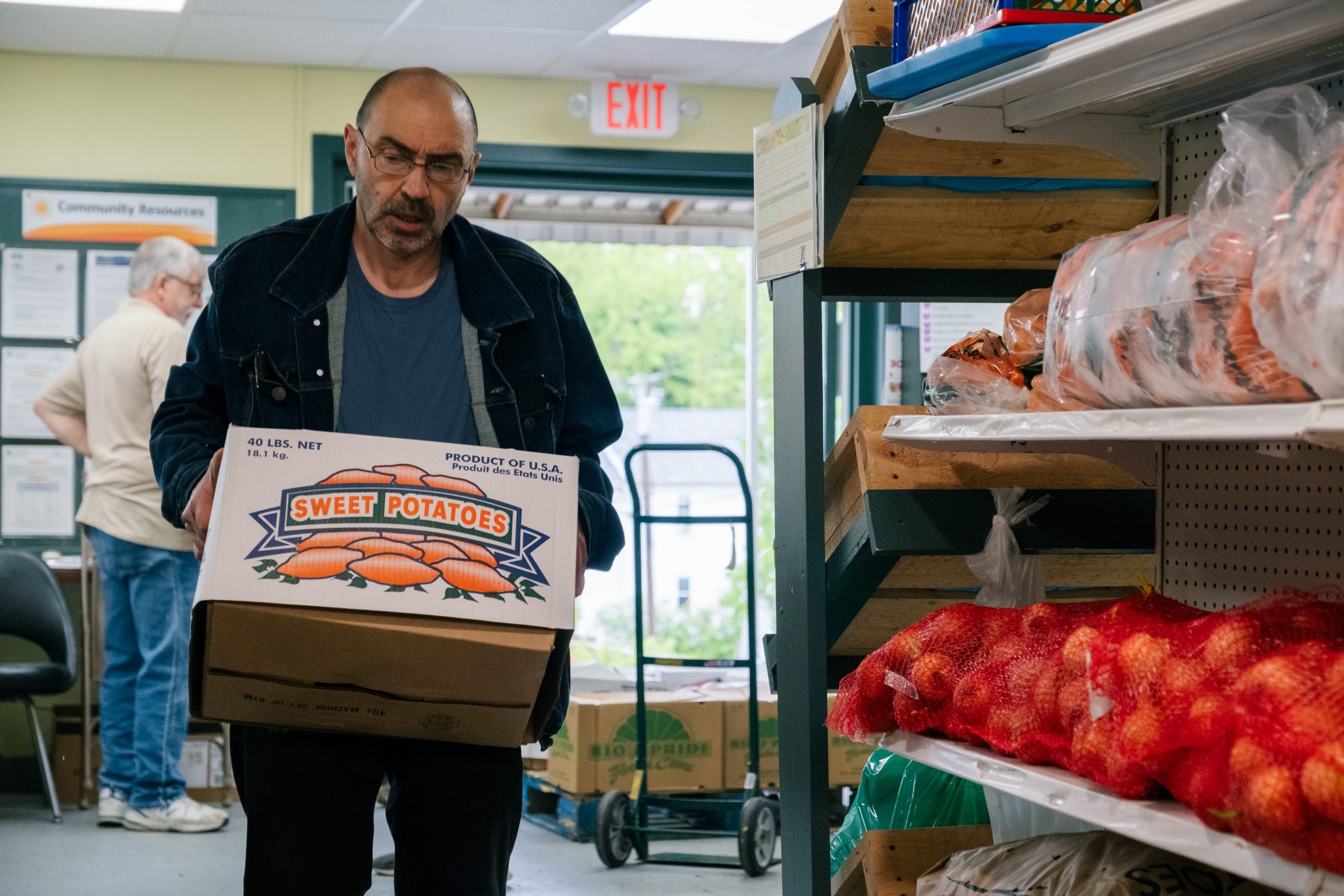 A man carries a box of food near shelves of produce.