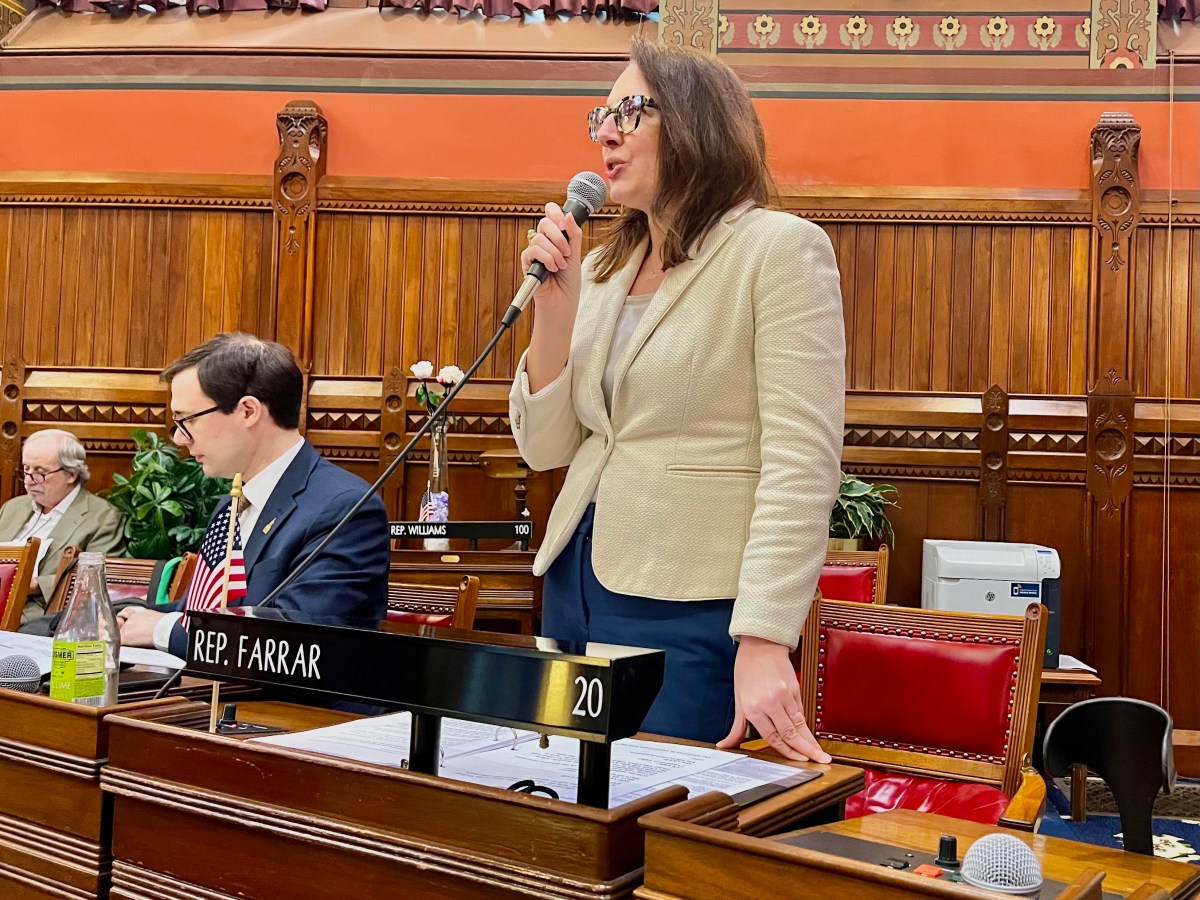 A woman in a white blazer stands in front of a chair and speaks into a microphone.