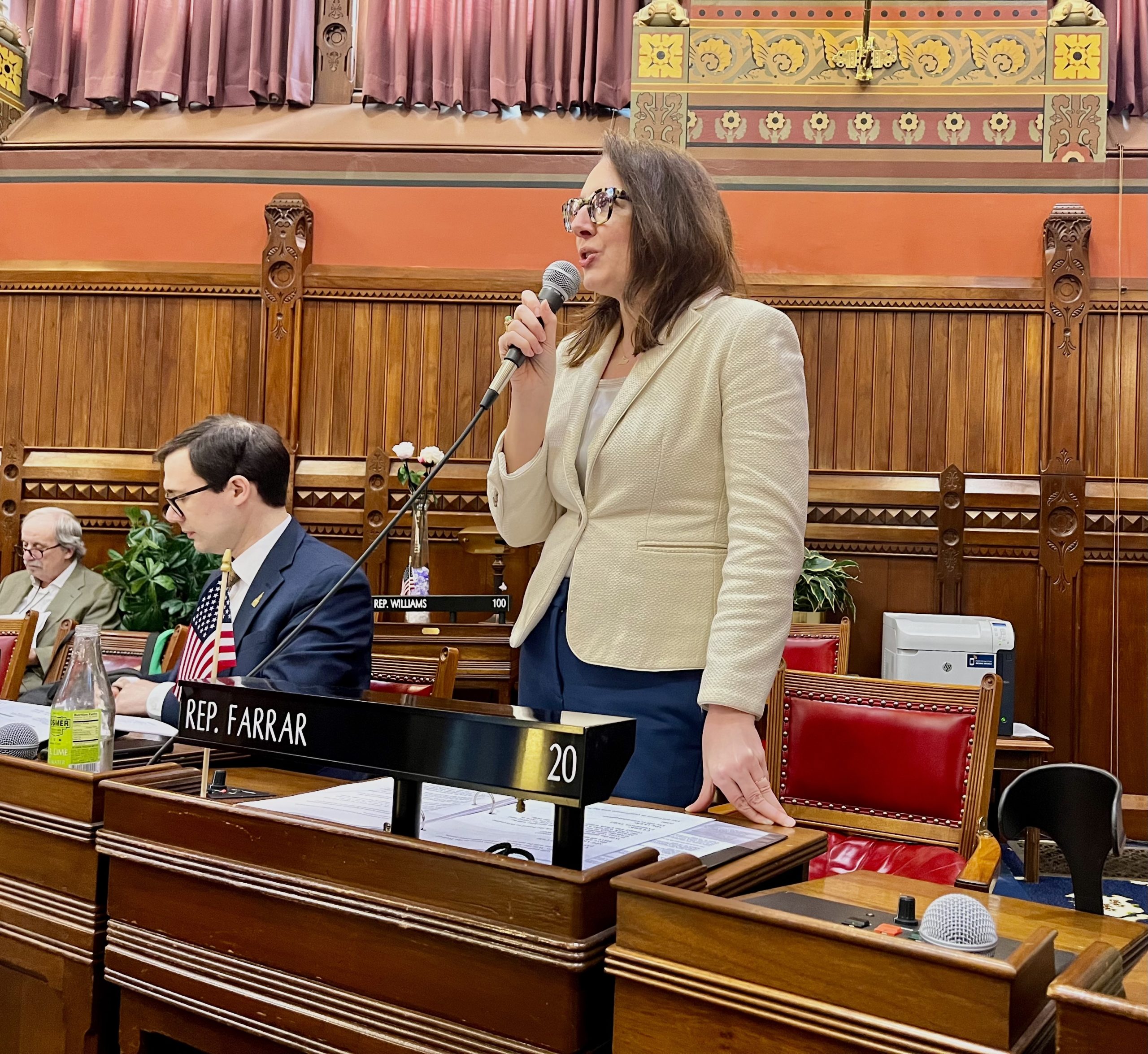A woman in a white blazer stands in front of a chair and speaks into a microphone.