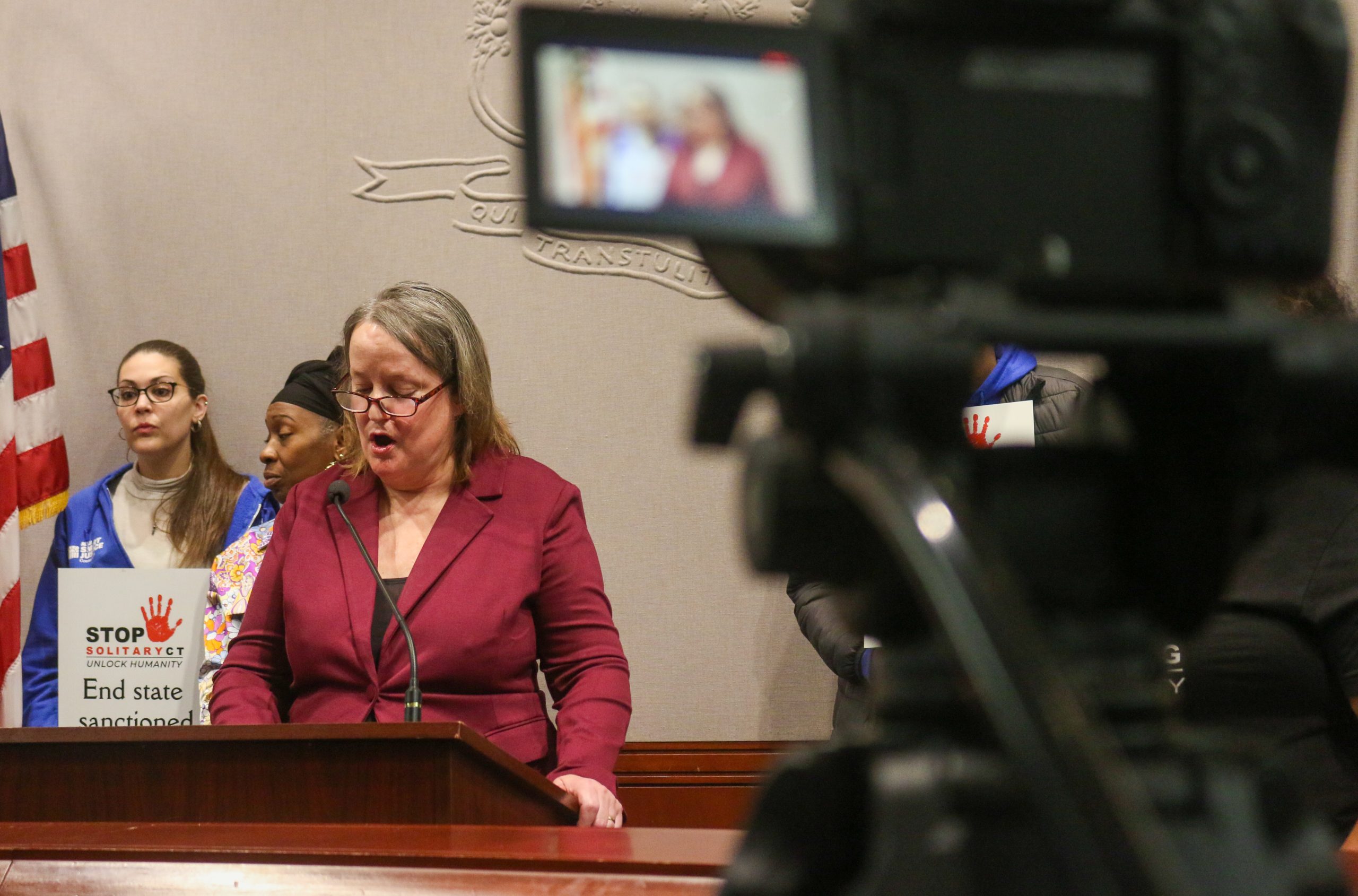Hilary Carpenter, standing in front of a podium with a microphone, speaks to a room full of people while two people are seen standing behind her.
