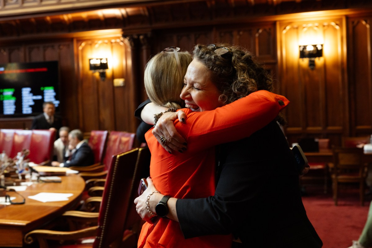 Two women embrace in a wood-paneled room as men sit at a table behind them.