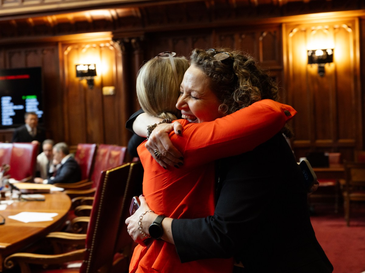 Two women embrace in a wood-paneled room as men sit at a table behind them.