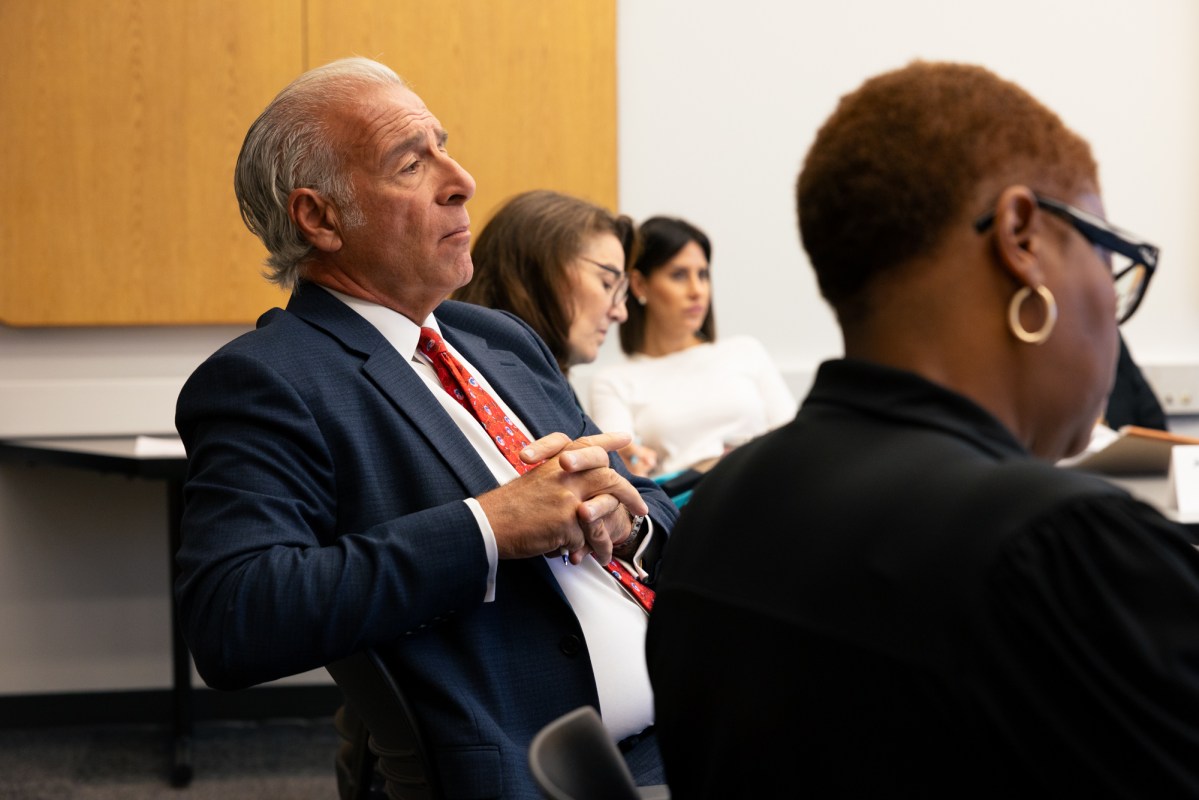 Tony Guerrera, a man with slicked-back white hair wearing a blue suit and red tie, leans back in his chair in a room with folding tables and wood panels on the walls. Other people sit in a circle around the table.
