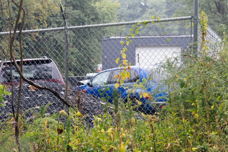 Two cars sit behind a chain-link fence and overgrown weeds.